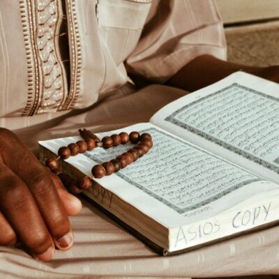 Crop anonymous religious African monk in robe reading holy Islamic book while sitting with rosary beads and praying