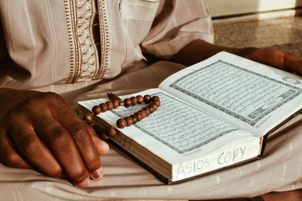 Crop anonymous religious African monk in robe reading holy Islamic book while sitting with rosary beads and praying